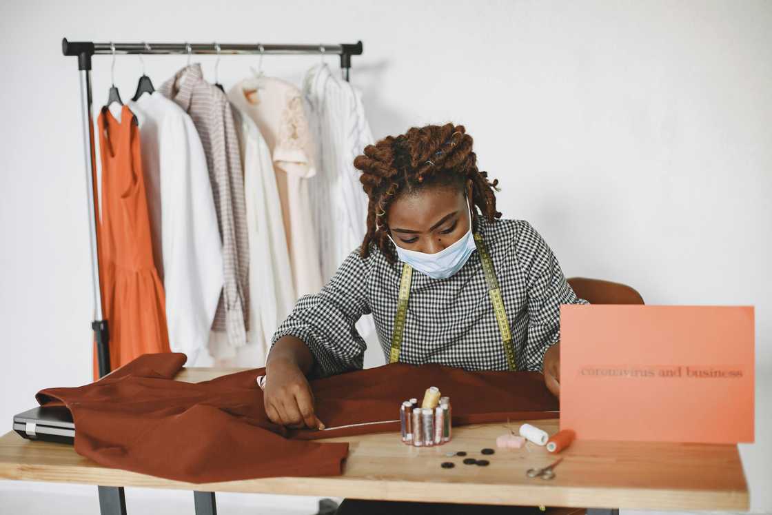 A seamstress in a black and white top and a face mask is sewing an outfit