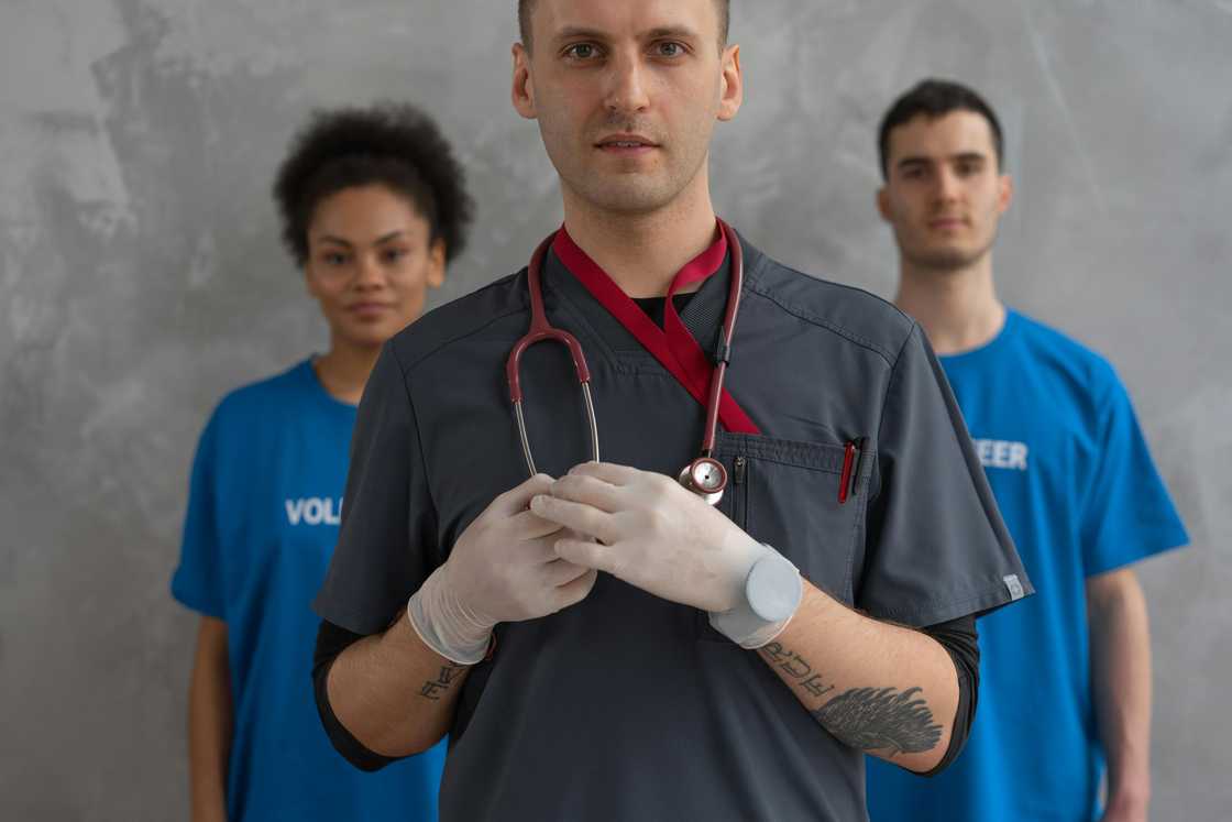 A man in a charcoal grey scrub suit standing in front a man and woman in nursing uniform