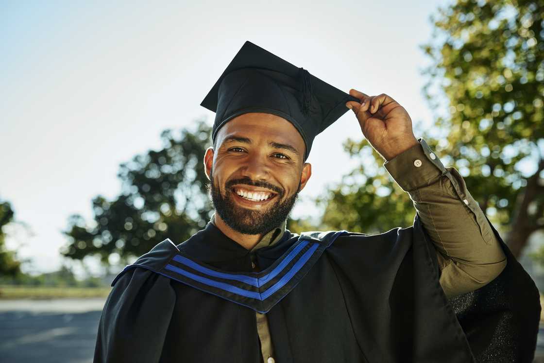 A young man smiles while holding his graduation cap