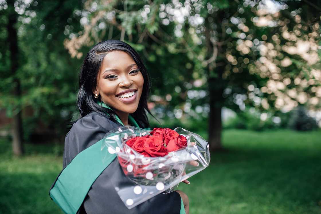 A lady in graduation gown standing in public park holding bouquet of red roses