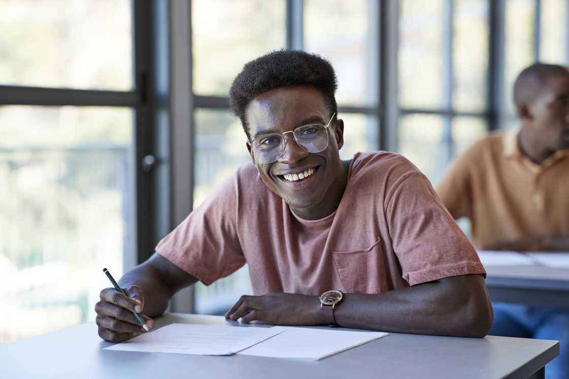 Smiling young male student sitting with exam papers at desk in community college classroom.